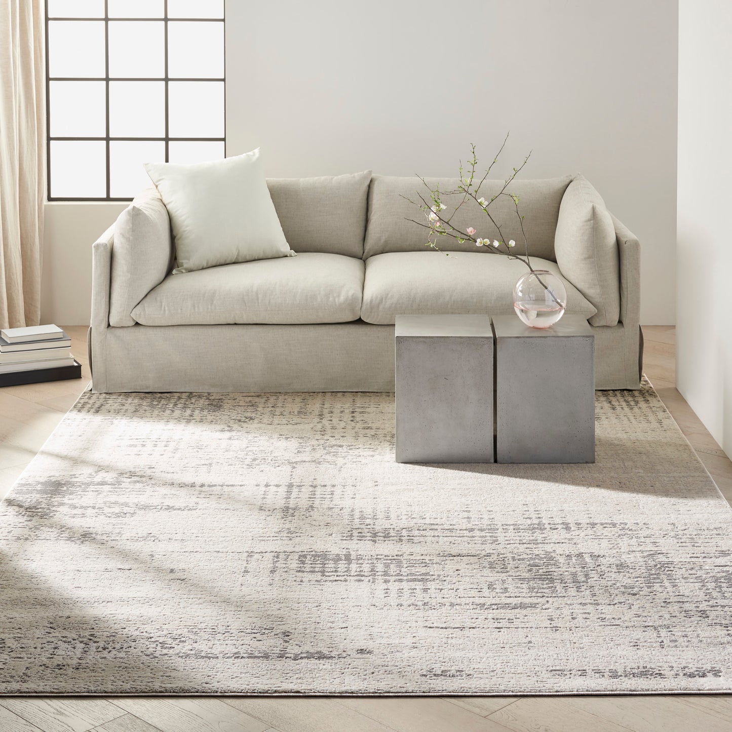 Neutral living room with beige fabric sofa, concrete cube tables, glass vase with blossoms, soft patterned rug.