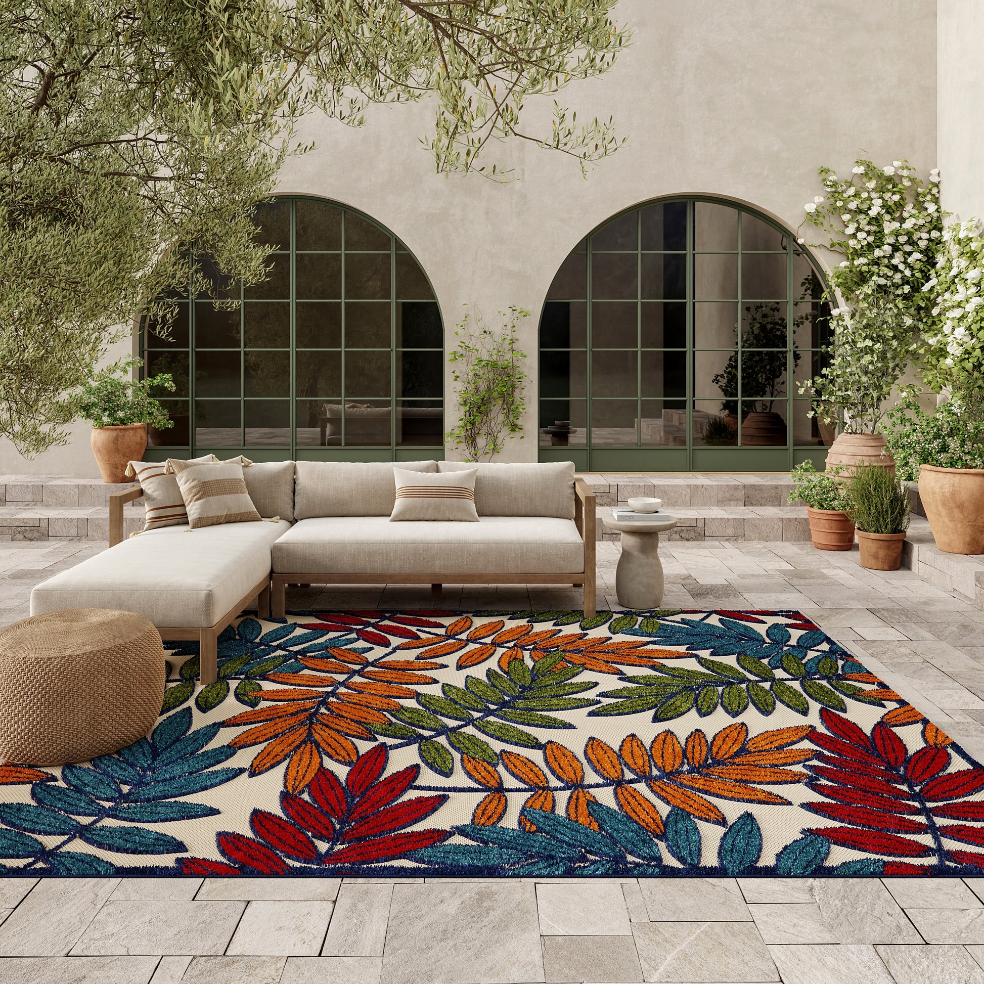 Outdoor patio with beige sectional sofa and a vibrant leaf-pattern rug in orange, blue, red, and green, plus potted plants.
