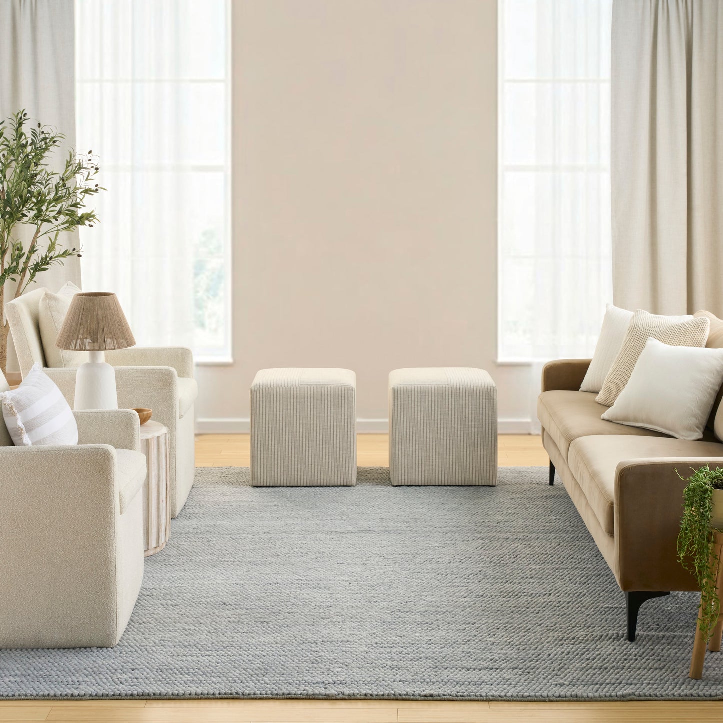 Neutral living room with cream upholstered armchairs, beige sofa, two square ottomans, gray rug, beige lamp, and potted plant.