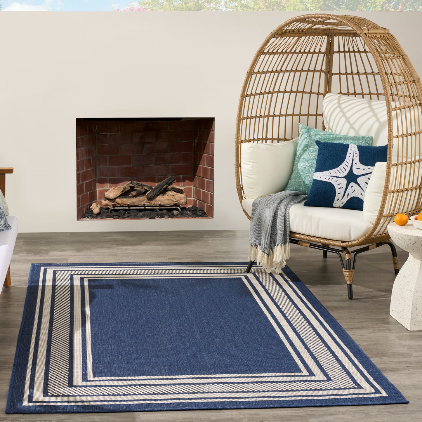 Blue rectangular rug with white border; round wicker egg chair with white cushions and teal starfish pillow beside fireplace.