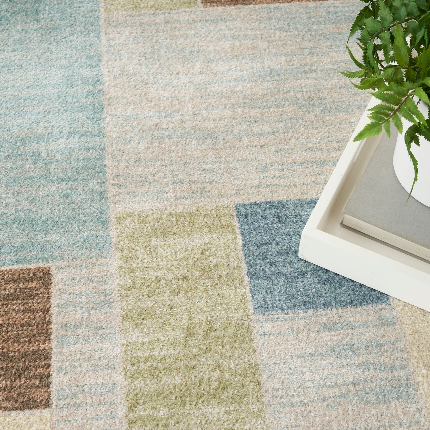 Geometric pastel area rug with color-block patches in blue, beige, green and brown; white tray and fern accent at edge.