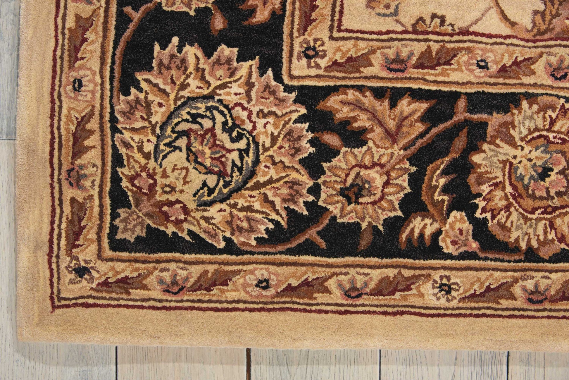 Ornate oriental rug corner with black field, gold-tan floral motifs, cream/beige border, laid on a wooden floor.