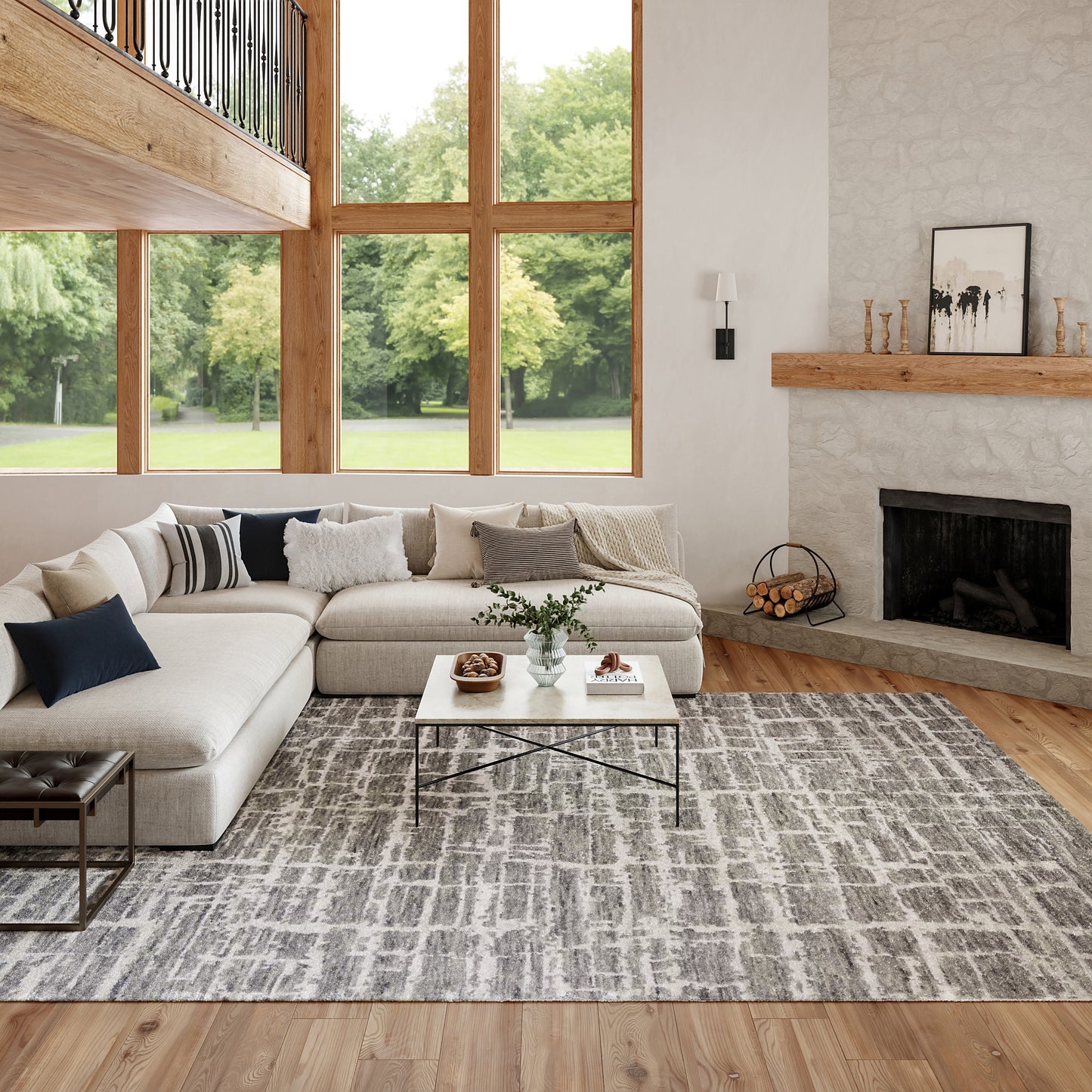 Neutral living room with a beige sectional, gray patterned rug, white coffee table, stone fireplace, and tall wood-framed windows.