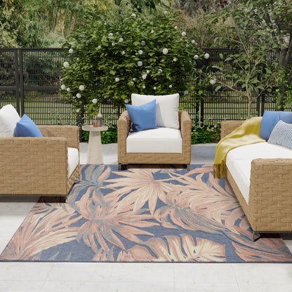Outdoor wicker patio set with white cushions, blue pillows, peach and blue palm-leaf rug, round side table, garden backdrop.