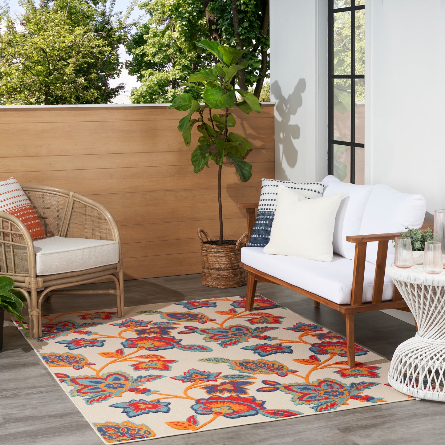Sunlit balcony with white-cushion sofa, rattan chair, floral outdoor rug, potted plant, wicker side table, wooden privacy screen.
