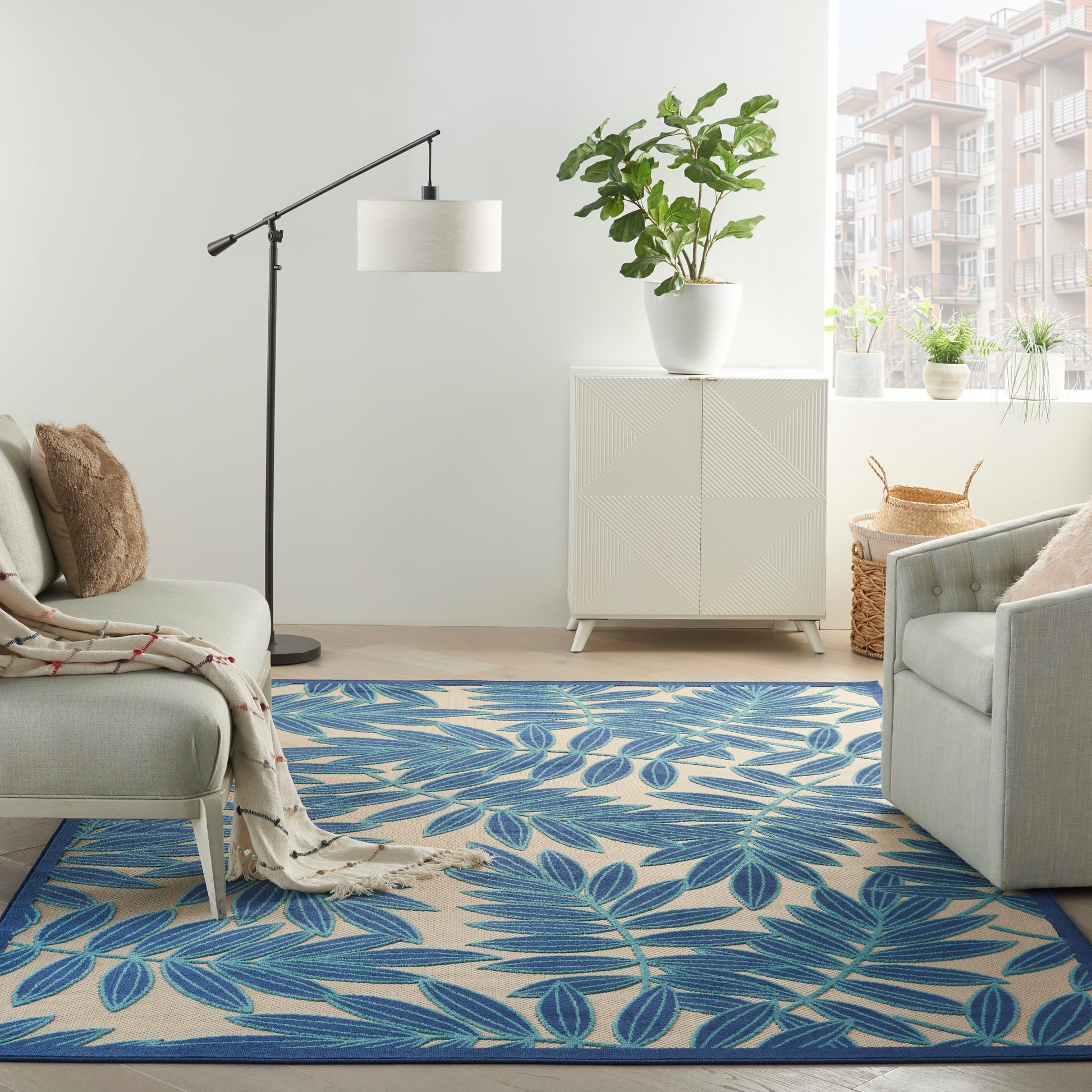 Modern living room with a blue leaf-pattern rug, light gray sofa & chair, white cabinet, potted plants.