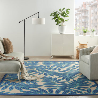 Modern living room with a blue leaf-pattern rug, light gray sofa & chair, white cabinet, potted plants.