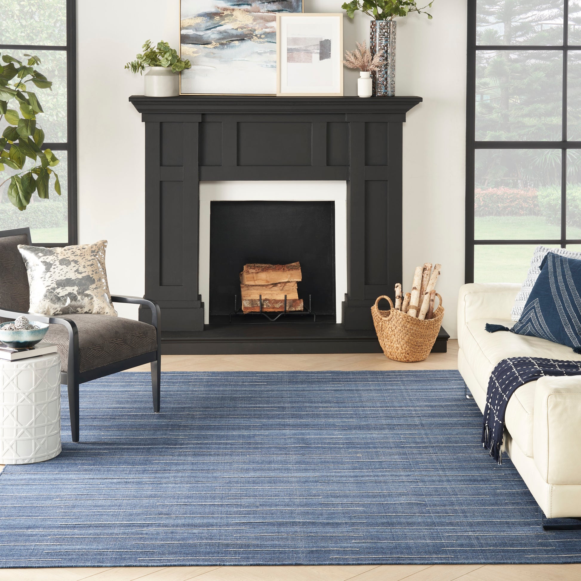 Blue rug on a living room floor with a cream sofa, gray chair, wicker basket, logs, and large windows.