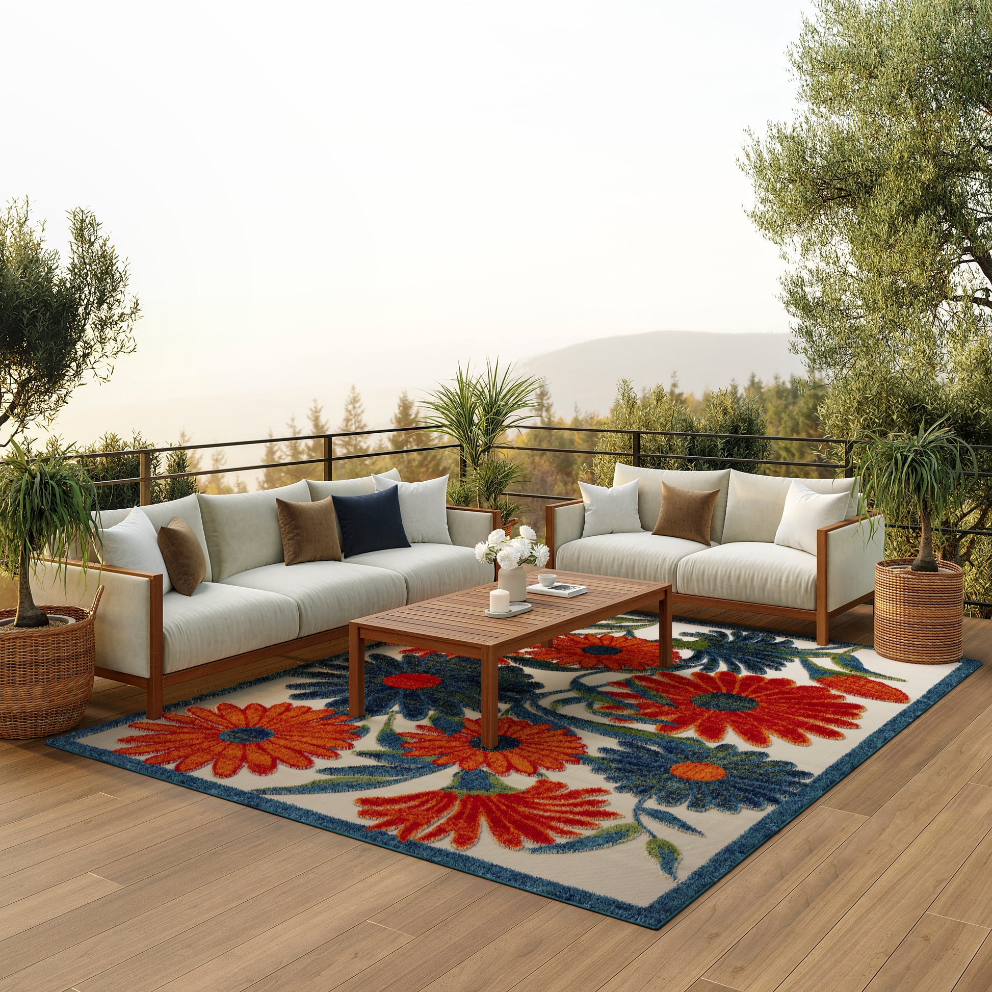 Outdoor lounge on a wooden deck with beige cushioned sectional, vibrant floral rug, coffee table and potted plants.