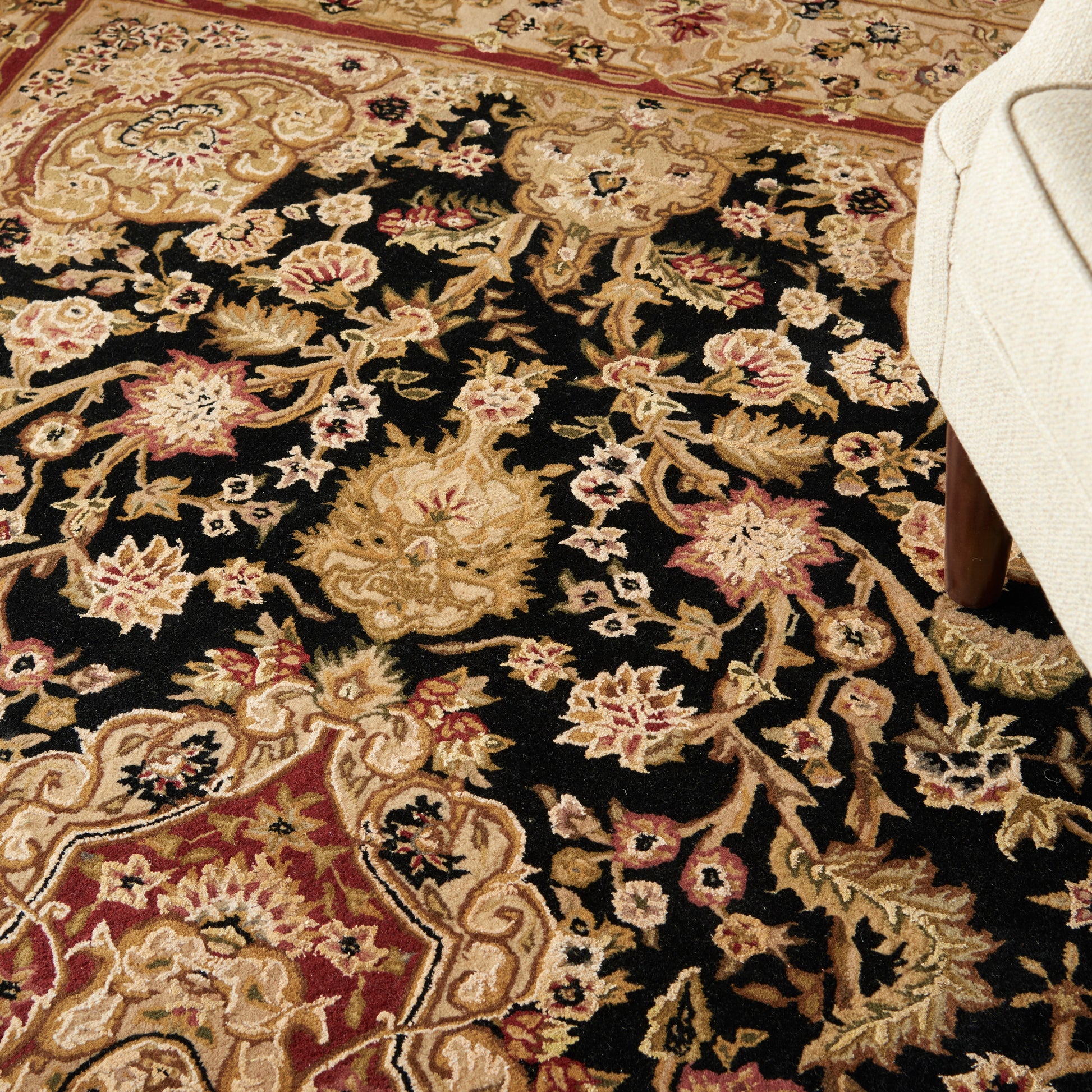 Intricate oriental rug with black field, gold and pink floral motifs, scrolling vines, beige border; edge of beige chair.
