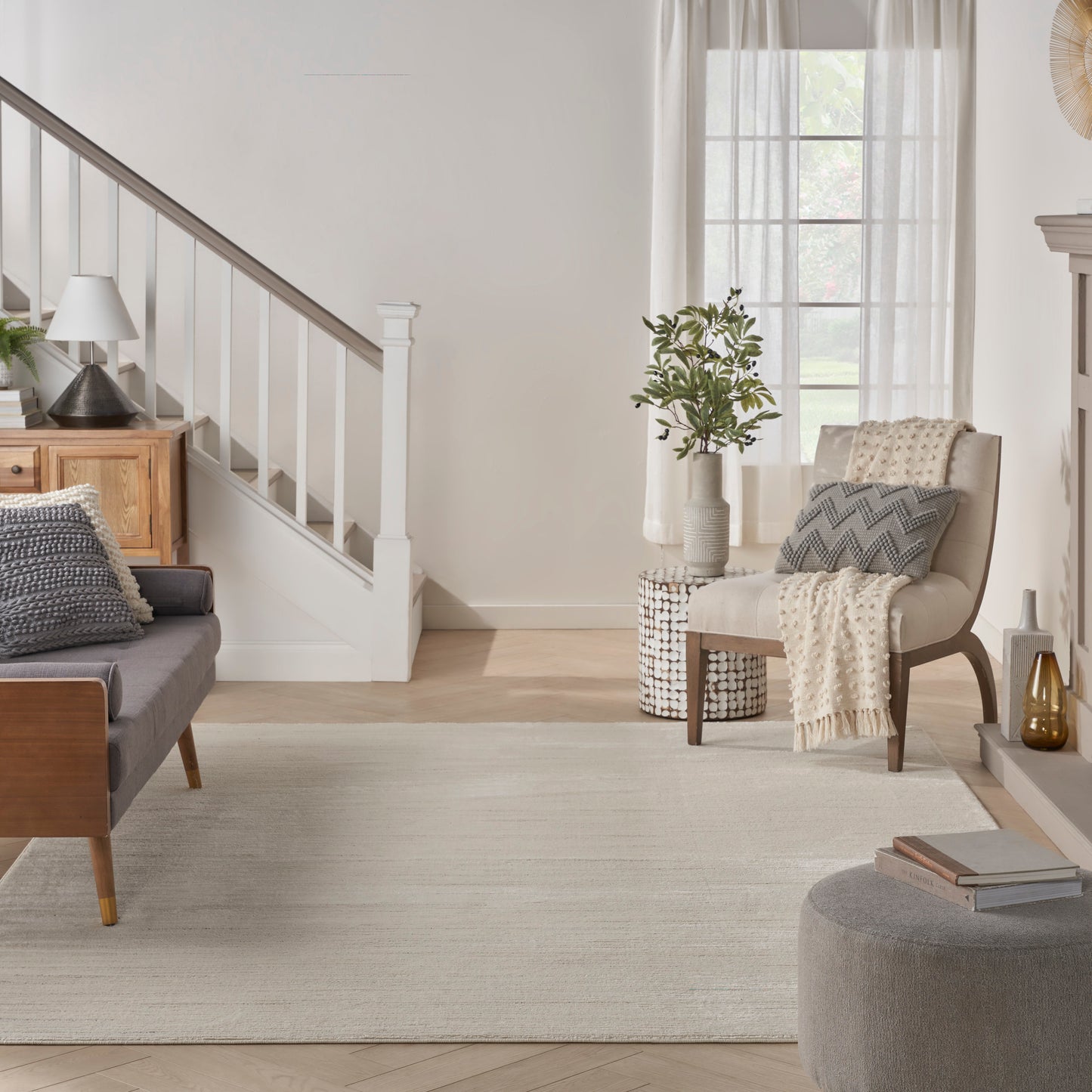 Neutral living room with light wood staircase, gray sofa, beige armchair, throw blanket, rug, and potted plant by window.