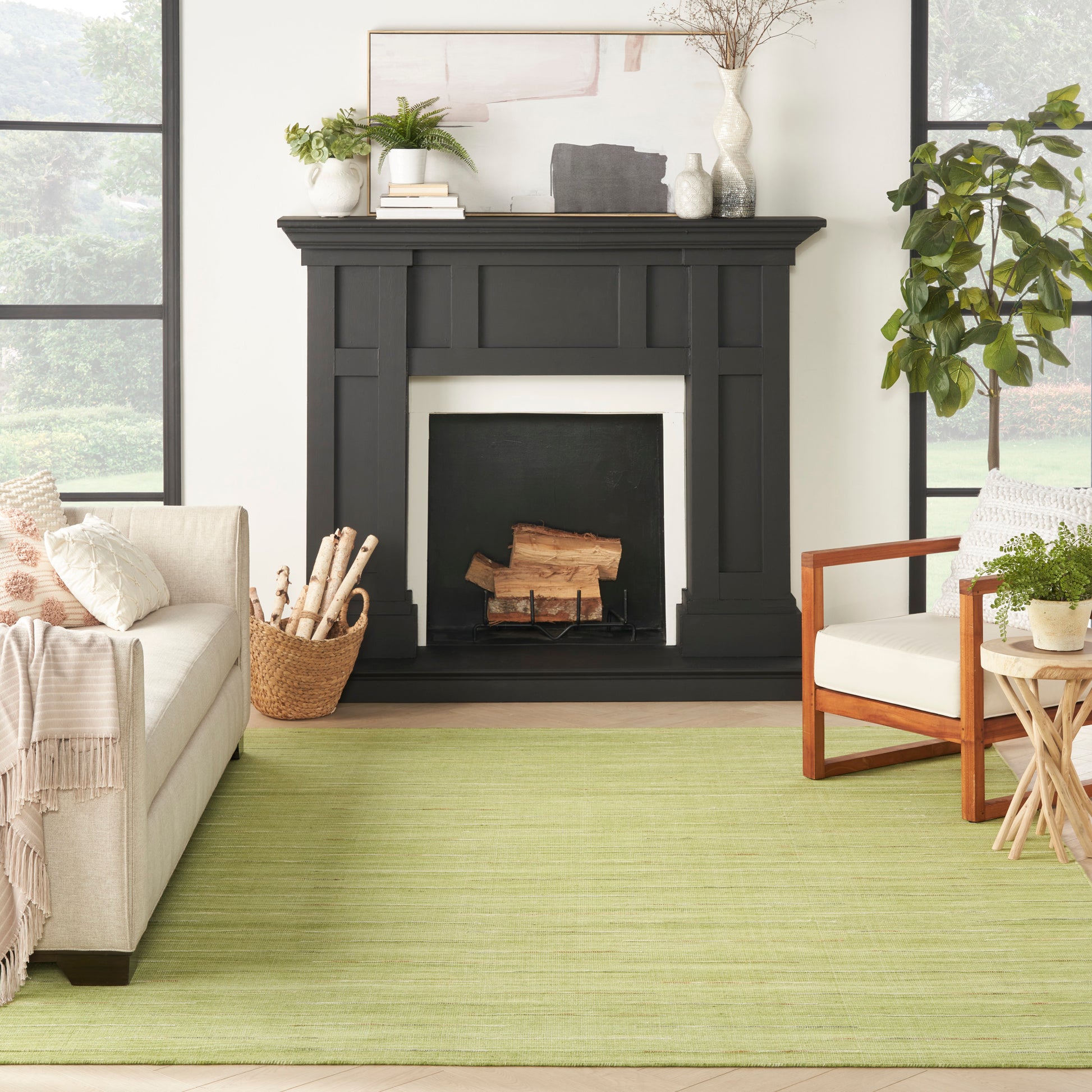 Living room with a black classical fireplace mantel, a green rug, beige sofa, wooden chair, and potted plants.