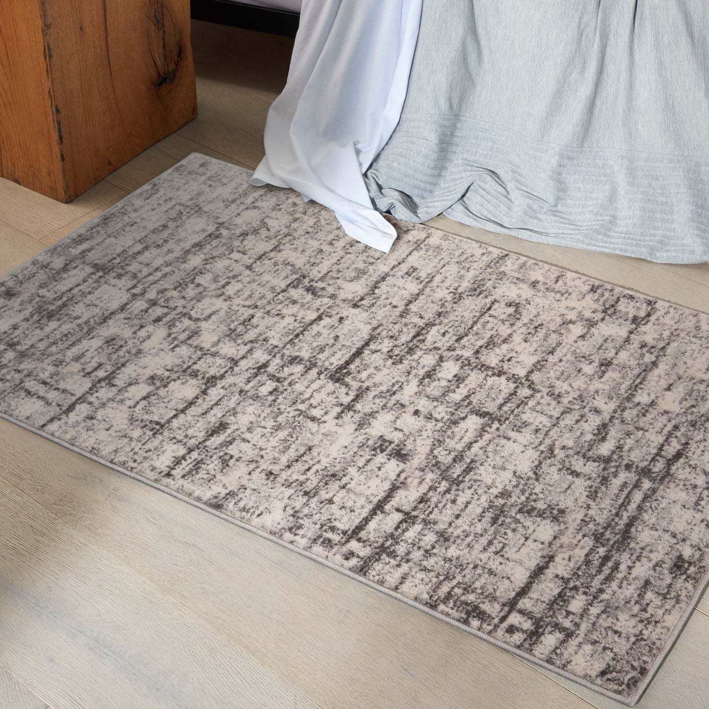 Rectangular beige-gray low-pile rug with irregular dark streaks on a light wood floor beside a bed.
