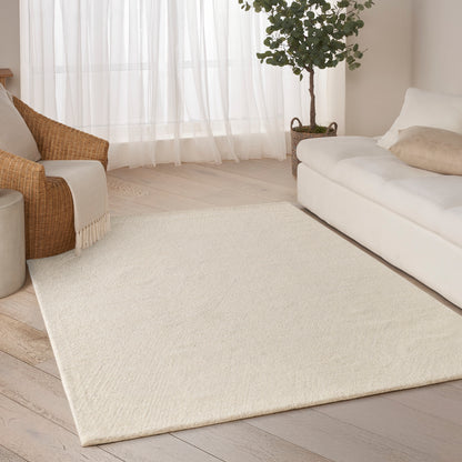 Soft cream rectangular area rug on pale wood floor in a bright living room with white sofa, wicker chair & potted tree.