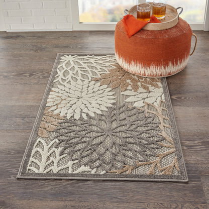 Rectangular outdoor rug with a large cream, taupe and brown botanical pattern on a gray border, beside an orange ottoman.