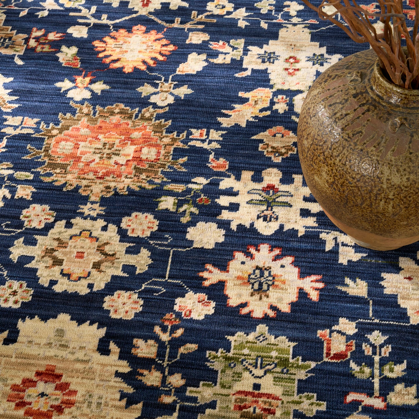 Blue oriental rug with cream, coral and green floral medallions; woven texture, brown ceramic vase on the right.