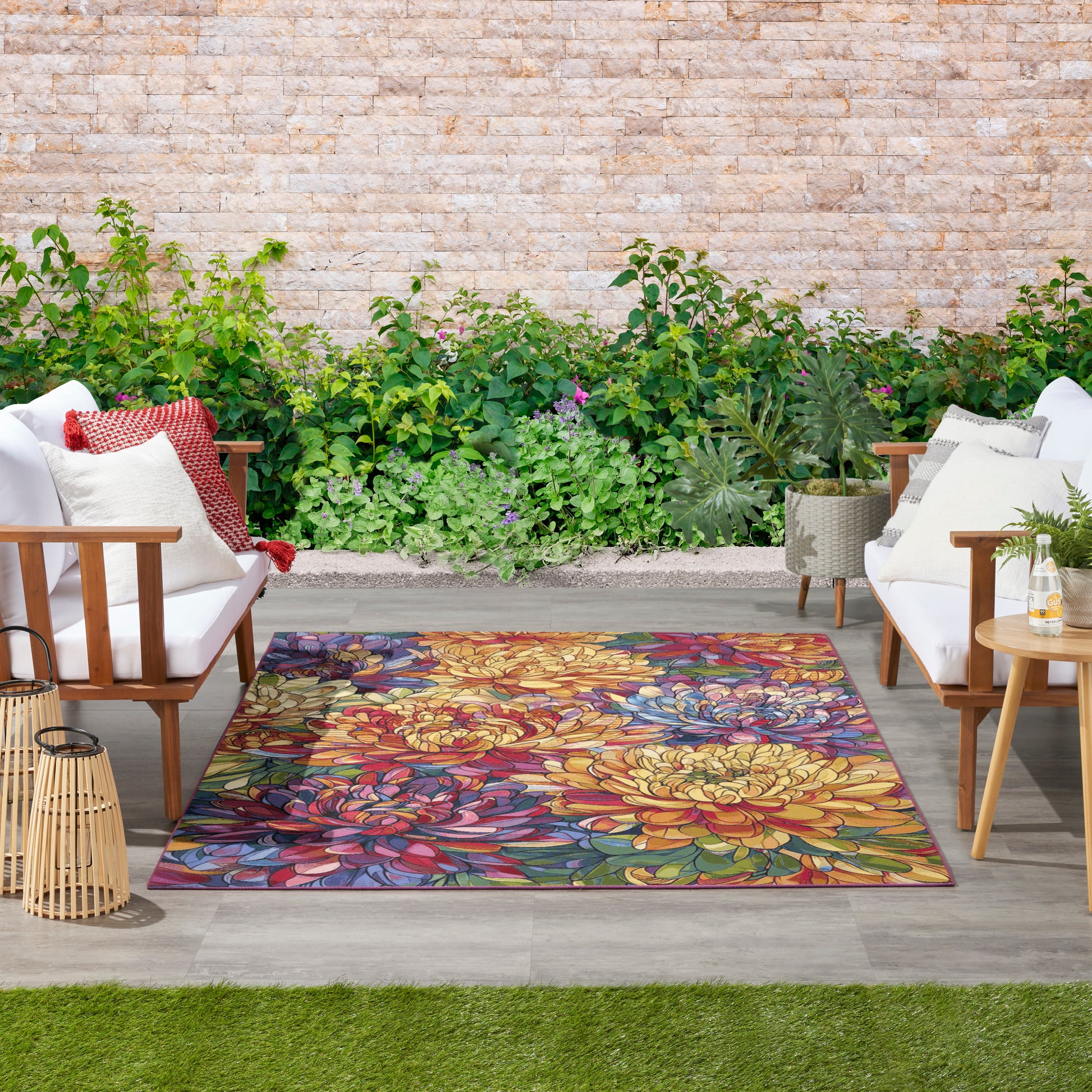 Vibrant multicolor floral outdoor rug framed by white cushioned wooden chairs on a stone patio with lush greenery.
