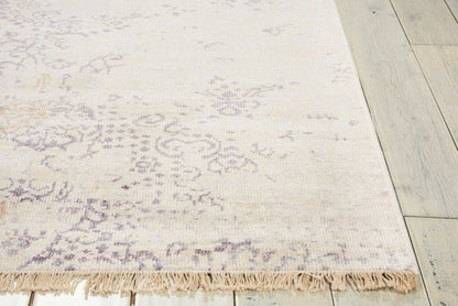 Cream area rug with subtle lavender and gray floral pattern and fringed edge on light wood floor.