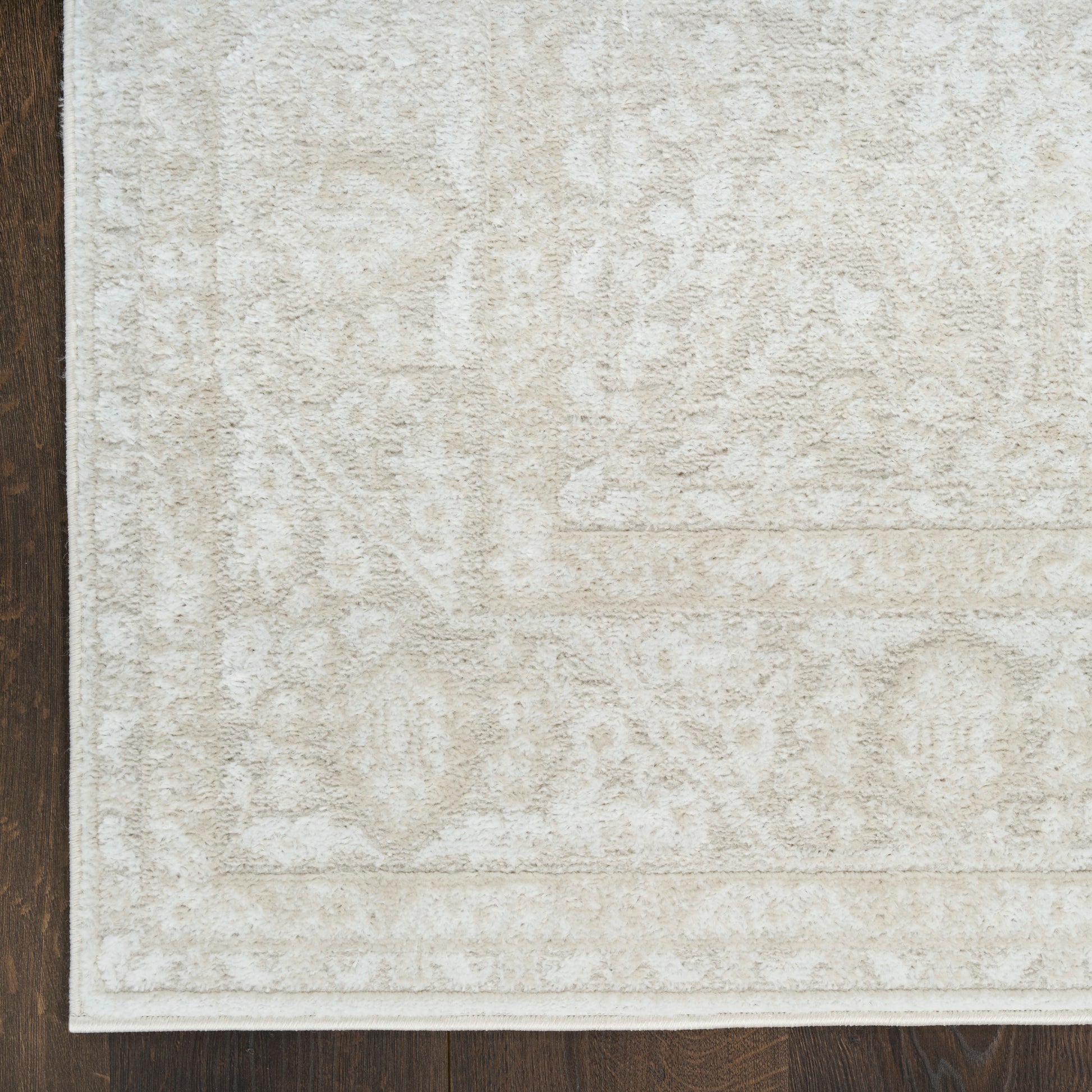 Beige area rug with subtle floral medallion pattern, light cream hues and white border on dark wood floor.