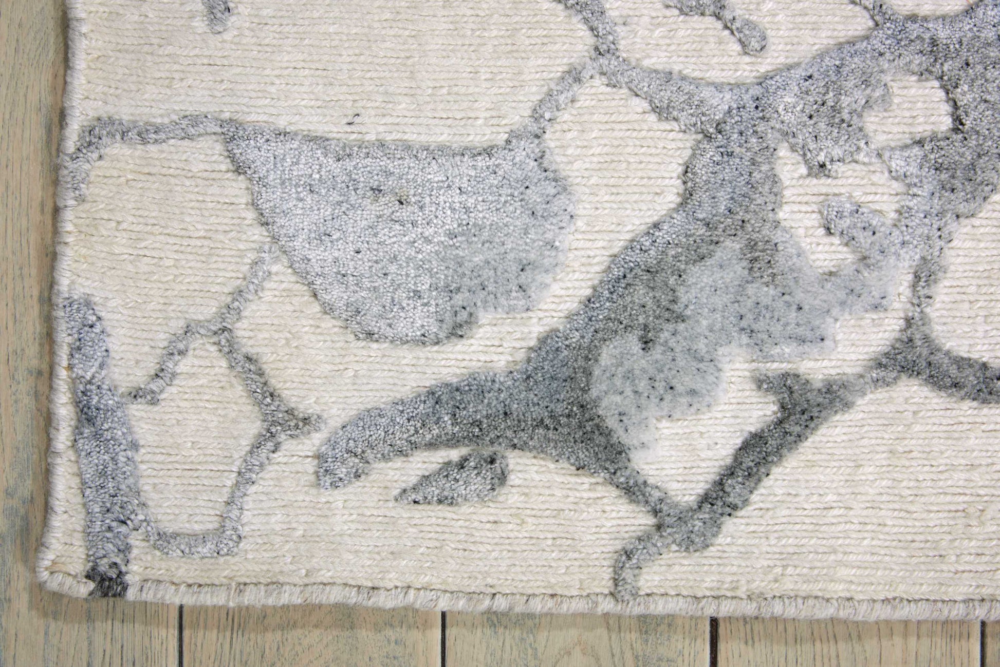 Cream rug with gray abstract vine/leaf pattern; textured pile, fringed edges on a wooden floor.