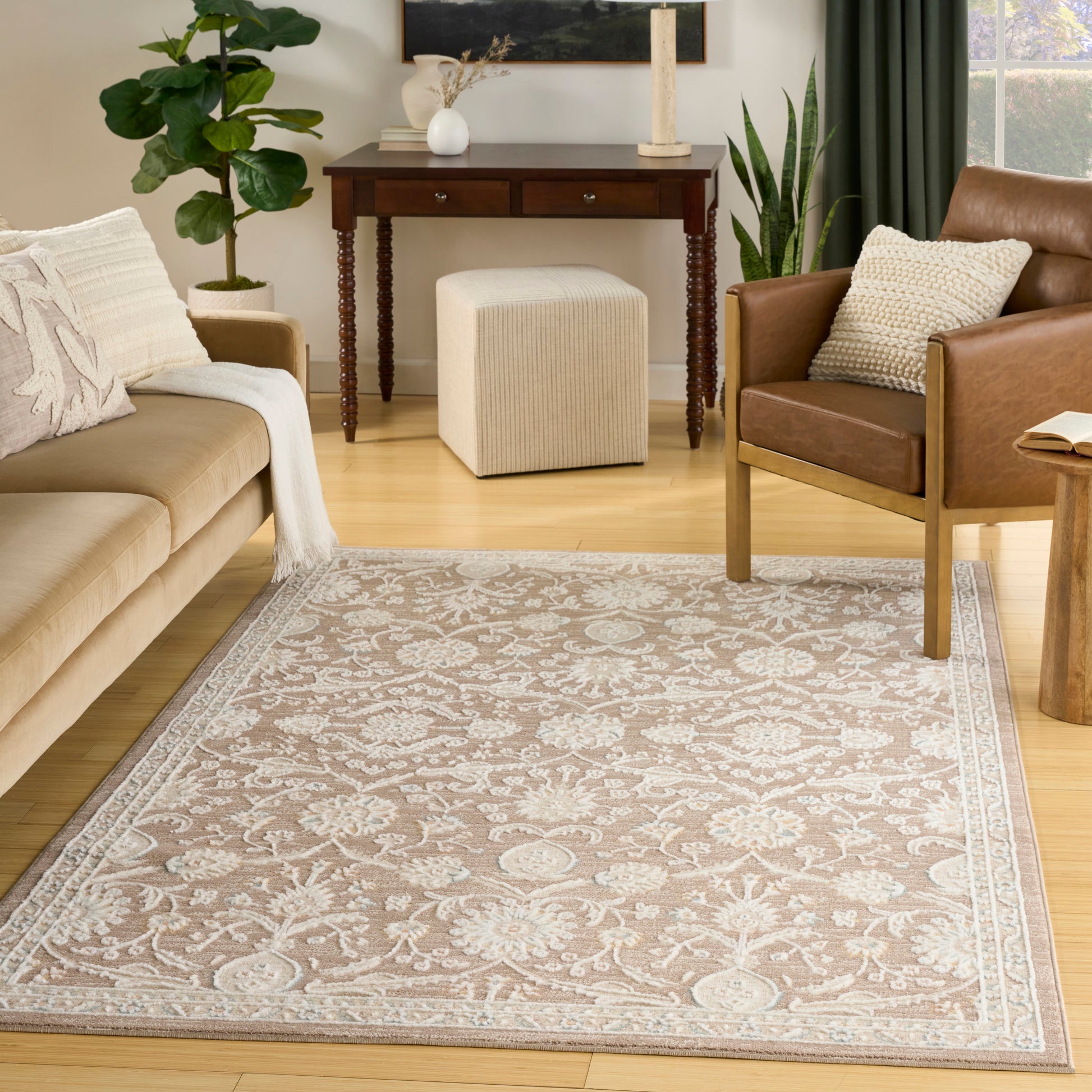 Beige floral area rug with ivory border in a cozy living room; tan sofa, brown leather chair, plants and a wooden console.