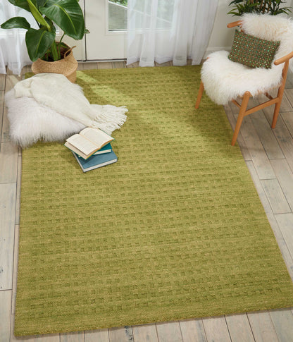 Olive-green textured area rug on light wood floor, with fluffy white throw, stack of books, potted plant, and wooden chair.