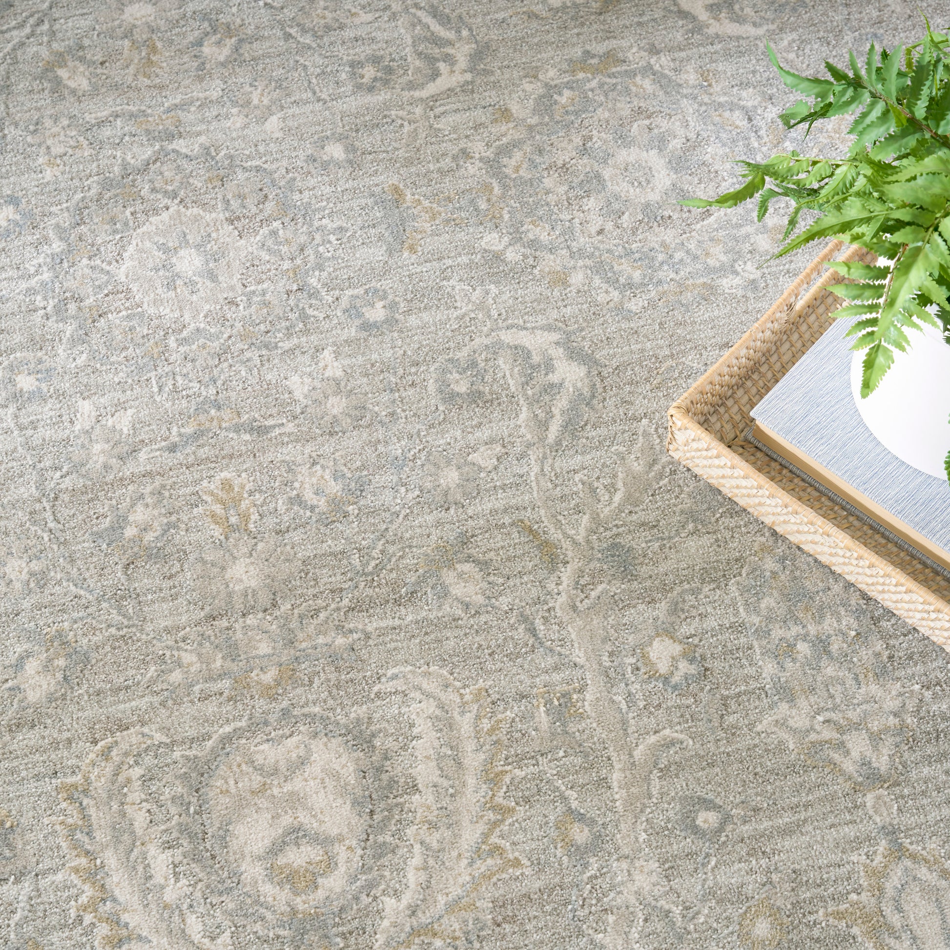 Light gray patterned area rug; wicker tray in corner holds a white pot with a green plant and a book.