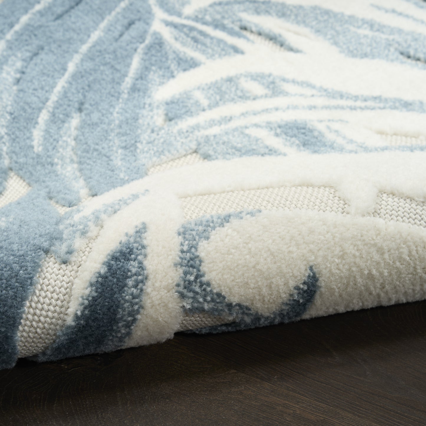 Blue and ivory plush rug with palm leaf pattern, folded edge shows soft pile and woven backing on dark wooden floor.