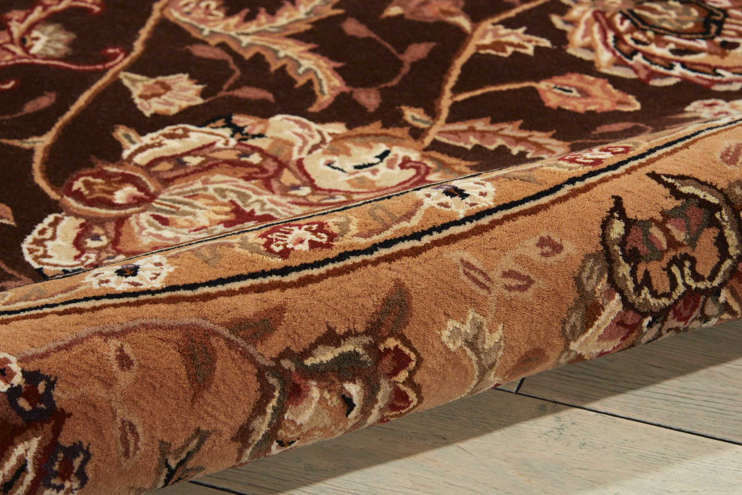 Close-up of a brown oriental rug with cream & red floral pattern & a rolled edge on a light wooden floor.