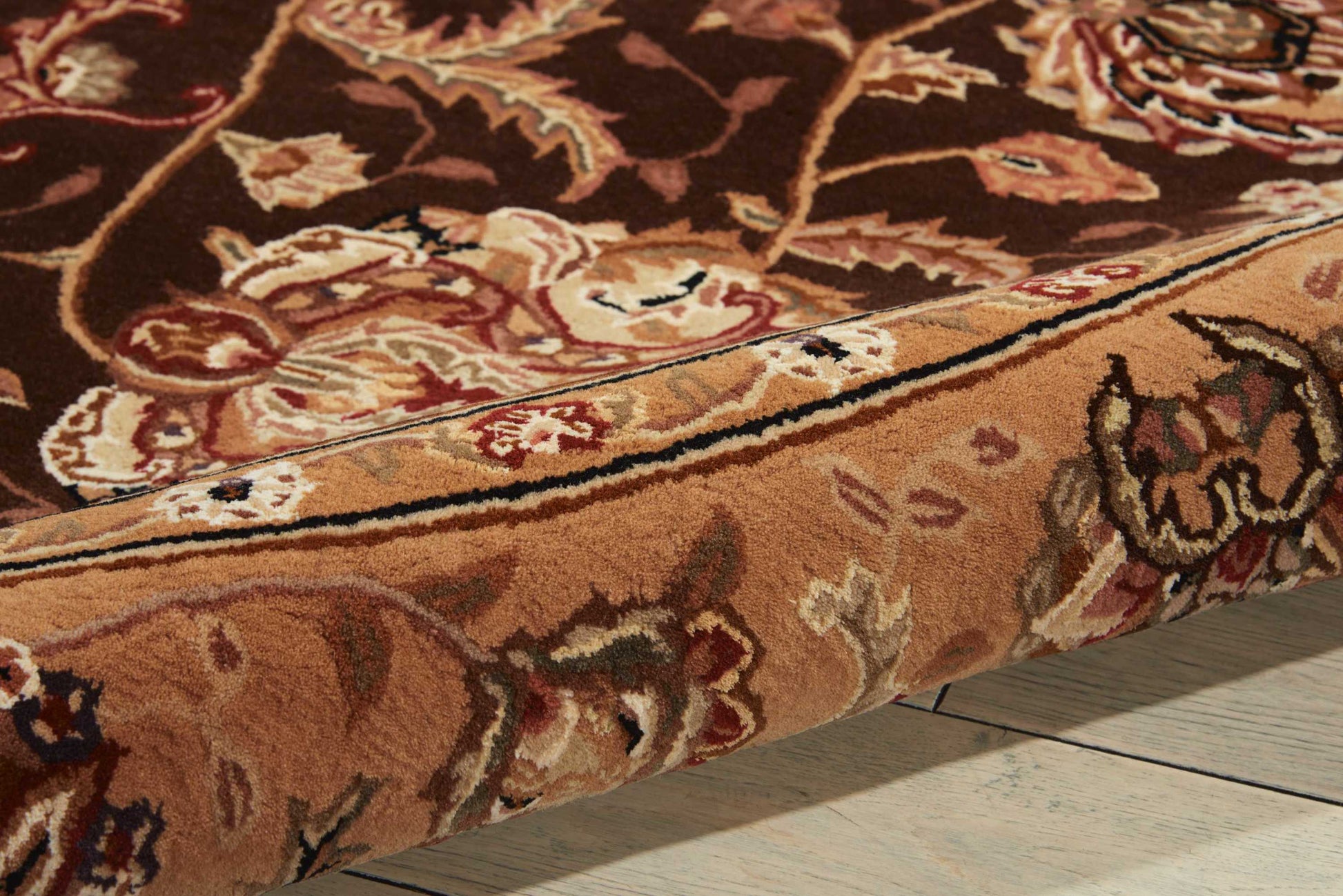 Close-up of a brown oriental rug with cream & red floral pattern & a rolled edge on a light wooden floor.