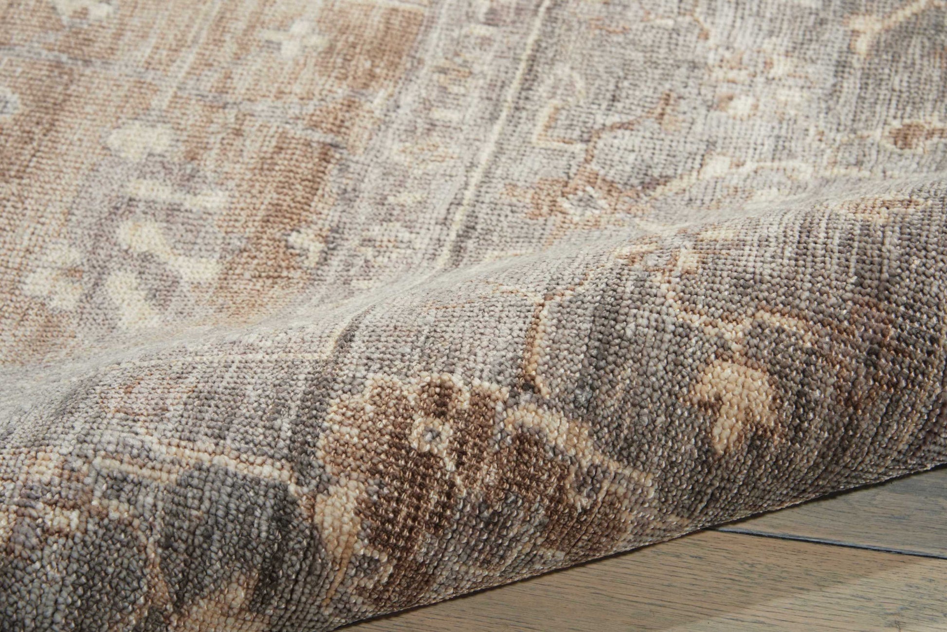 Muted beige-brown patterned area rug with floral motifs, partially rolled on a wooden floor.
