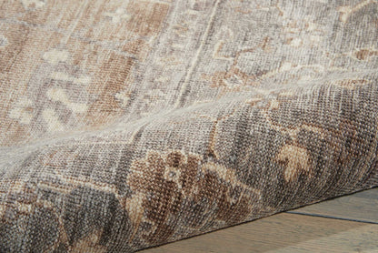 Muted beige-brown patterned area rug with floral motifs, partially rolled on a wooden floor.