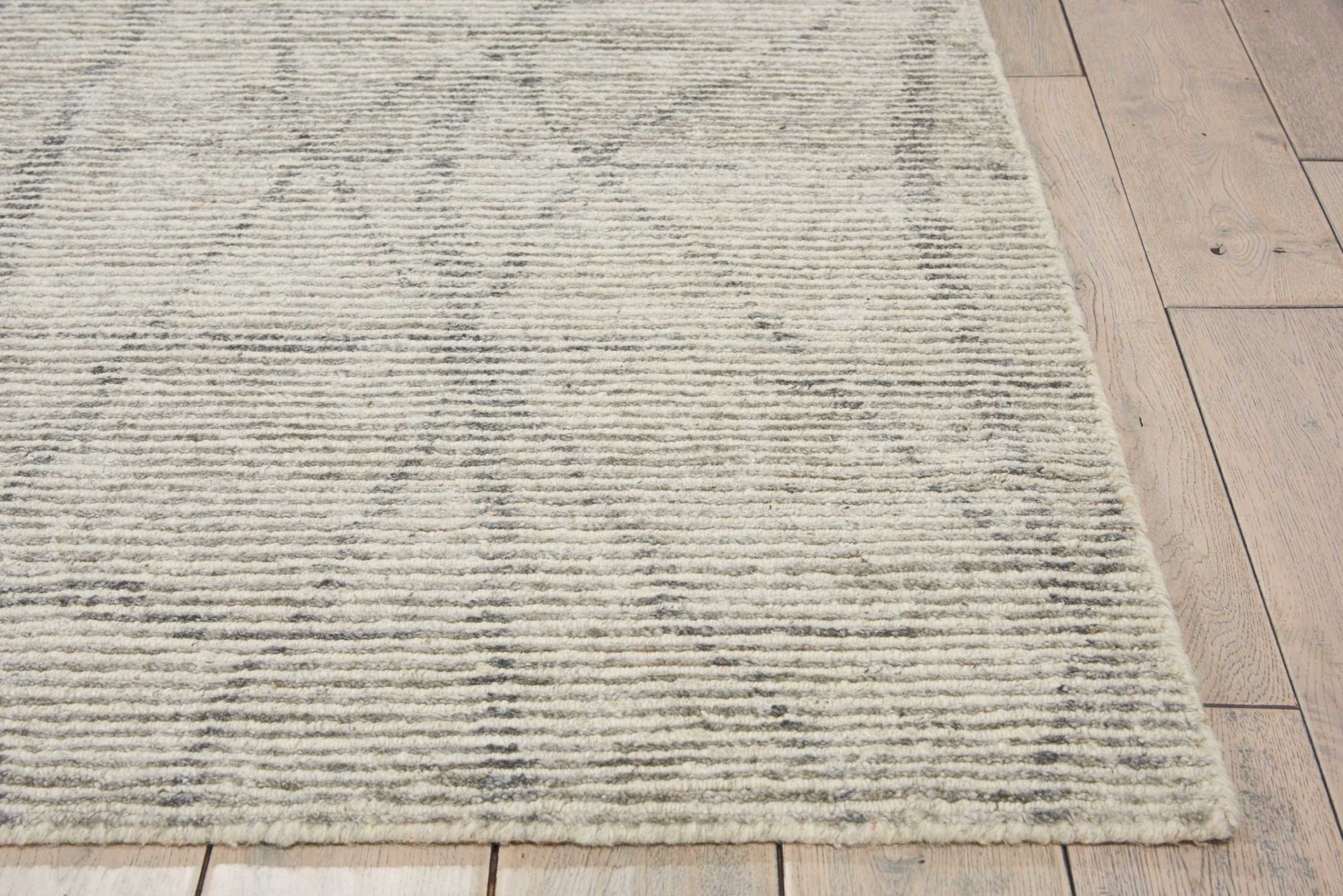 Beige rectangular area rug with subtle gray geometric motif and fringed border on light wood floor.