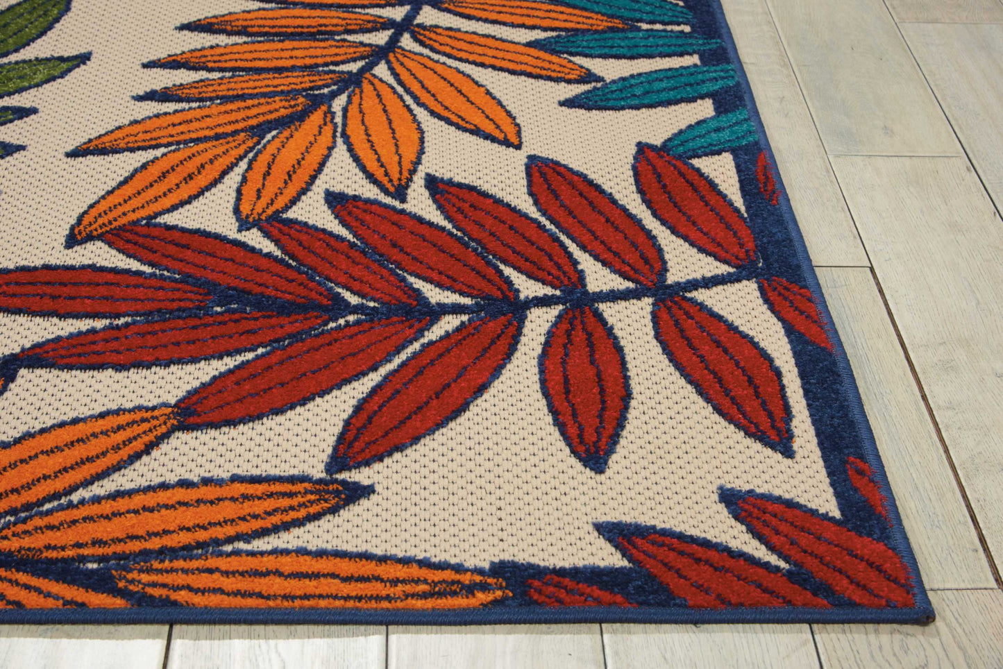Leaf-pattern rug featuring orange, red, & teal leaves on a beige background with a dark blue border, on a light wooden floor.