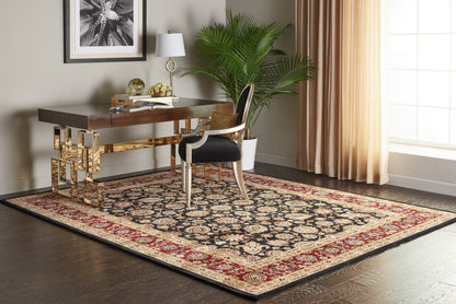 Home office with dark wood desk on an ornate red black beige rug, gold-accented legs, white lamp, plant, and black-and-white upholstered chair.