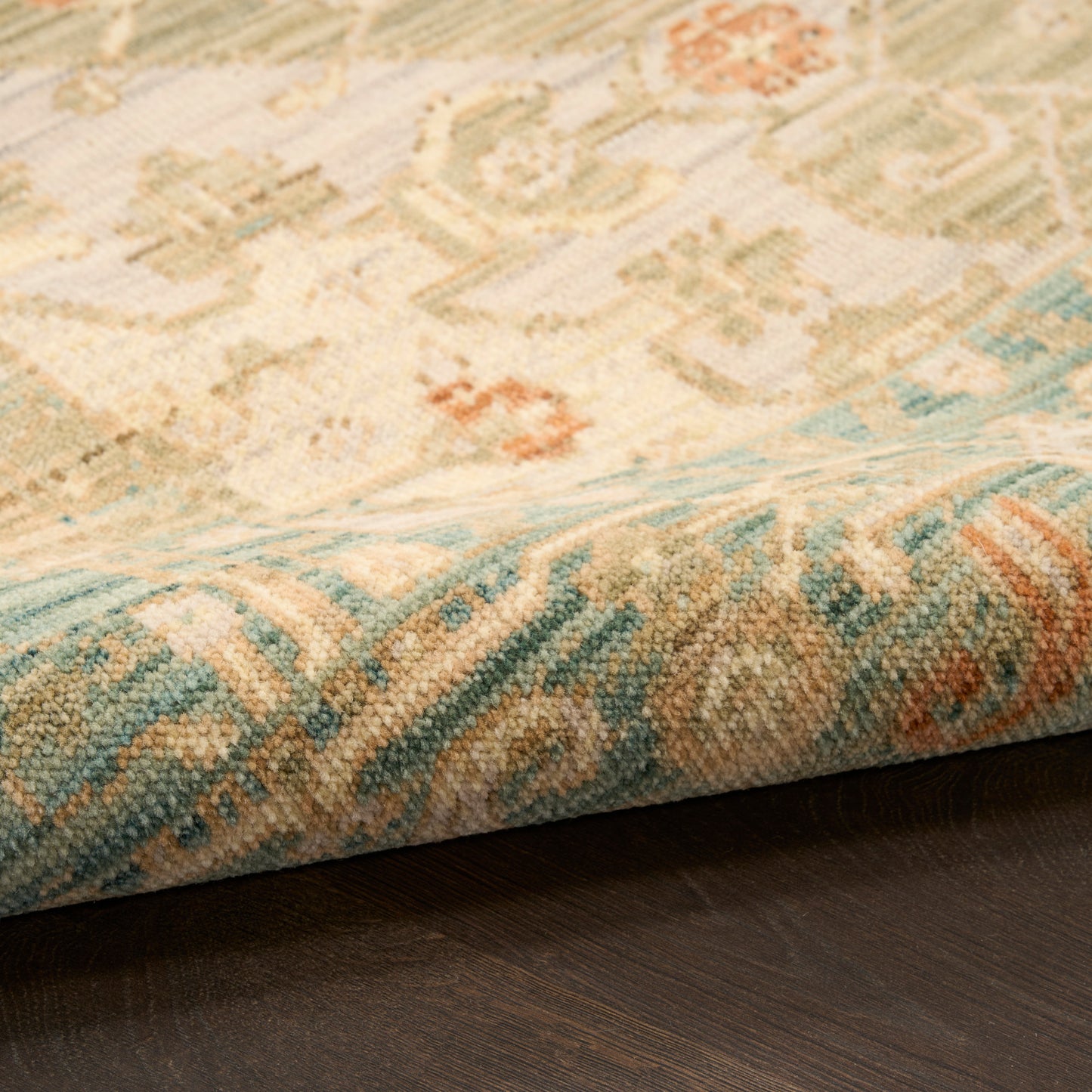 Pastel-toned oriental rug close-up with beige, blue & peach motifs and folded edge on dark wood floor.