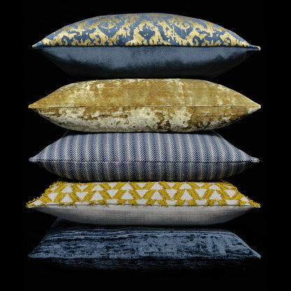 Five decorative throw pillows stacked: indigo with gold motif, gold velvet, blue herringbone, yellow triangles, navy velvet.