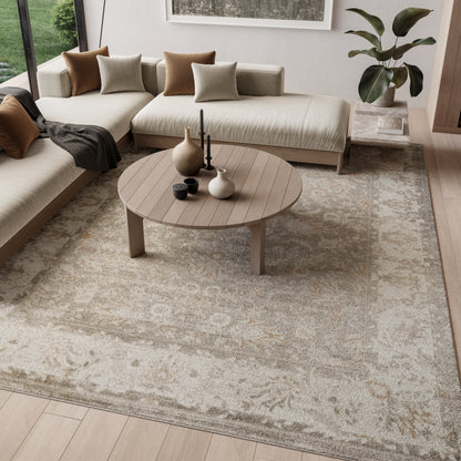 Round wooden coffee table on beige floral rug beside a neutral sectional with brown cushions and a potted plant.