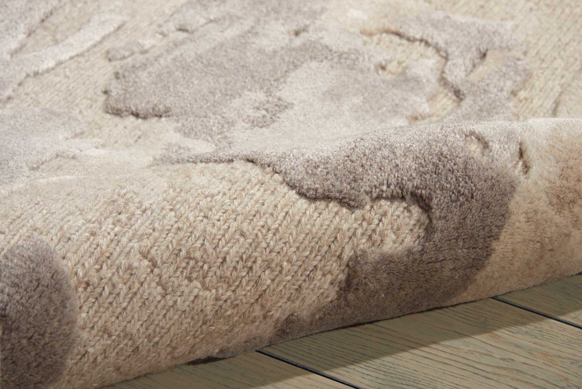 Beige and taupe patterned area rug with thick plush pile, rolled edge over light wood floor.