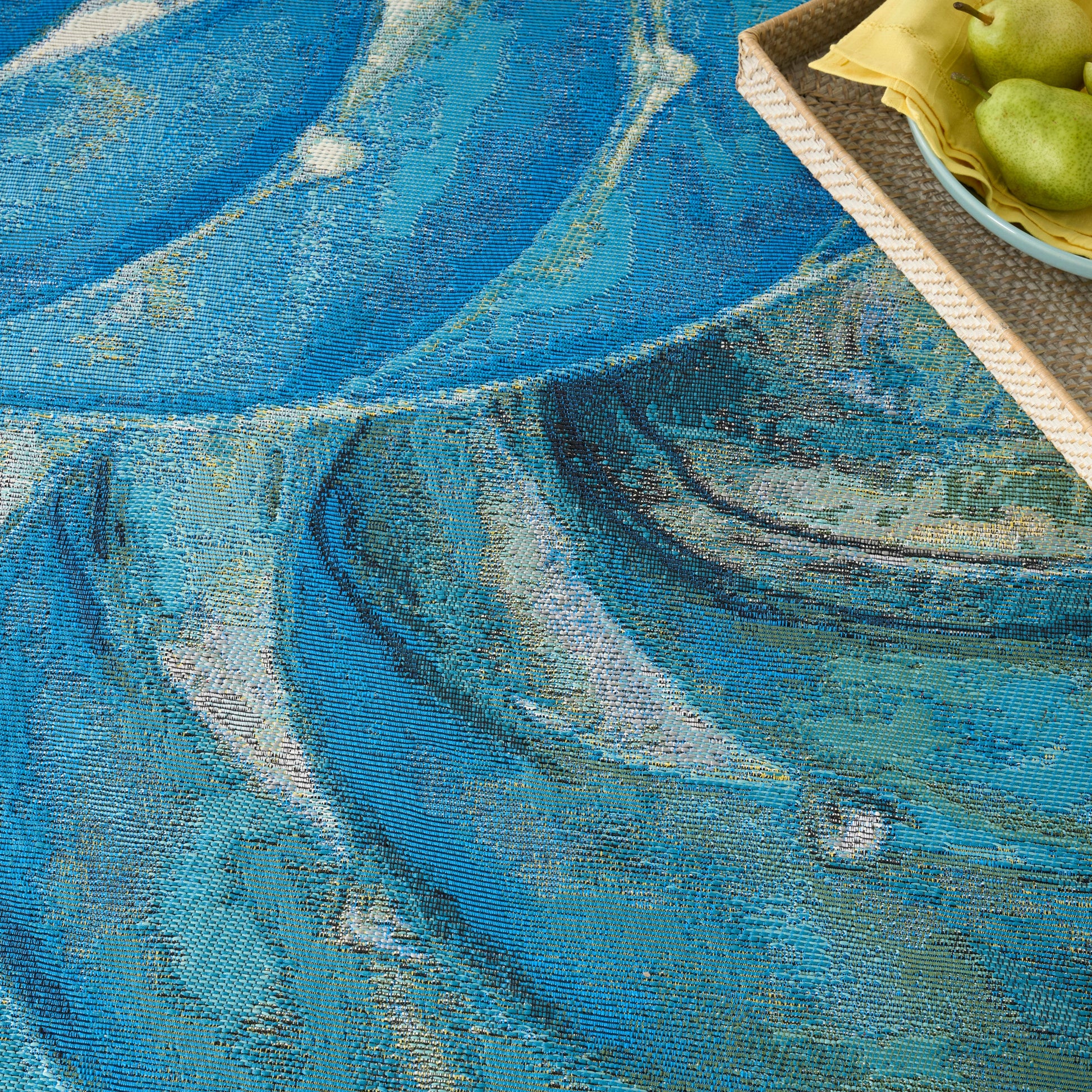 Blue abstract woven rug with teal swirls; woven texture; corner wicker tray with green pears and yellow napkin.