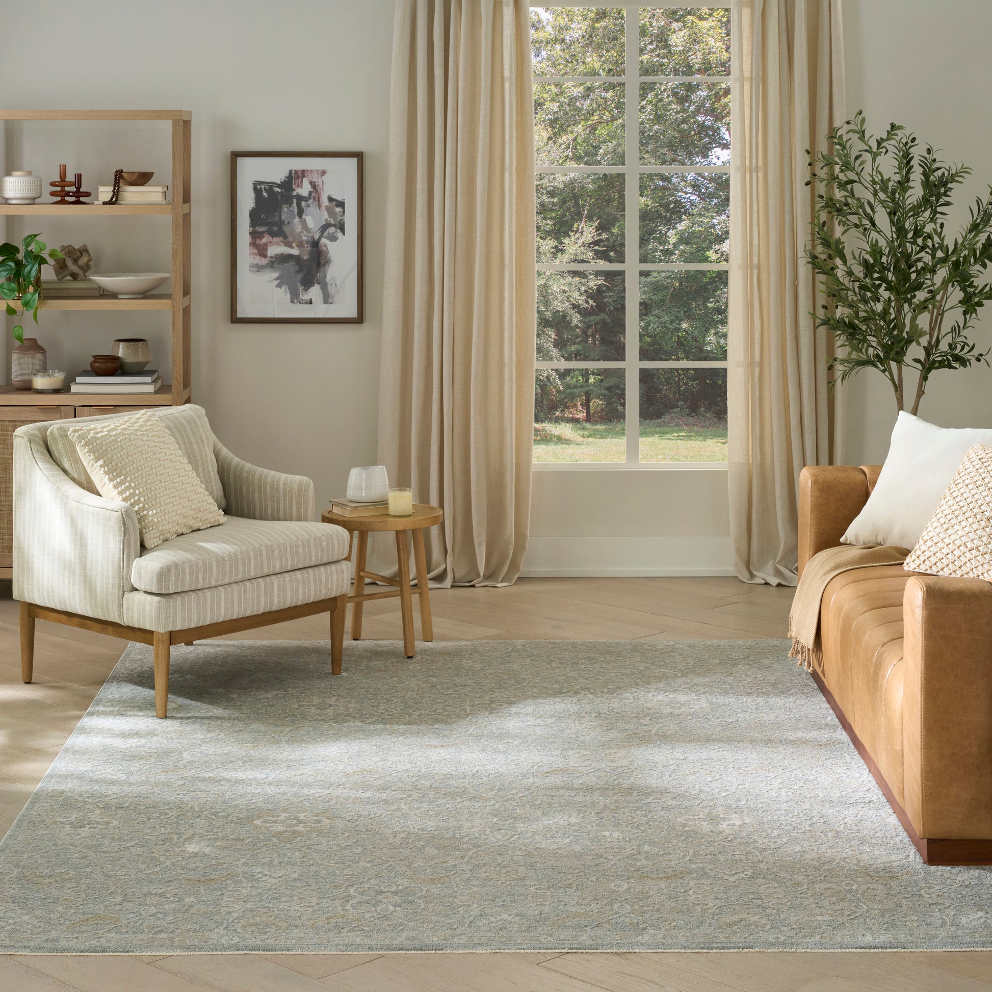 Bright living room with beige armchair, caramel sofa, neutral cushions, wooden shelf, framed art, grey rug, and large window.