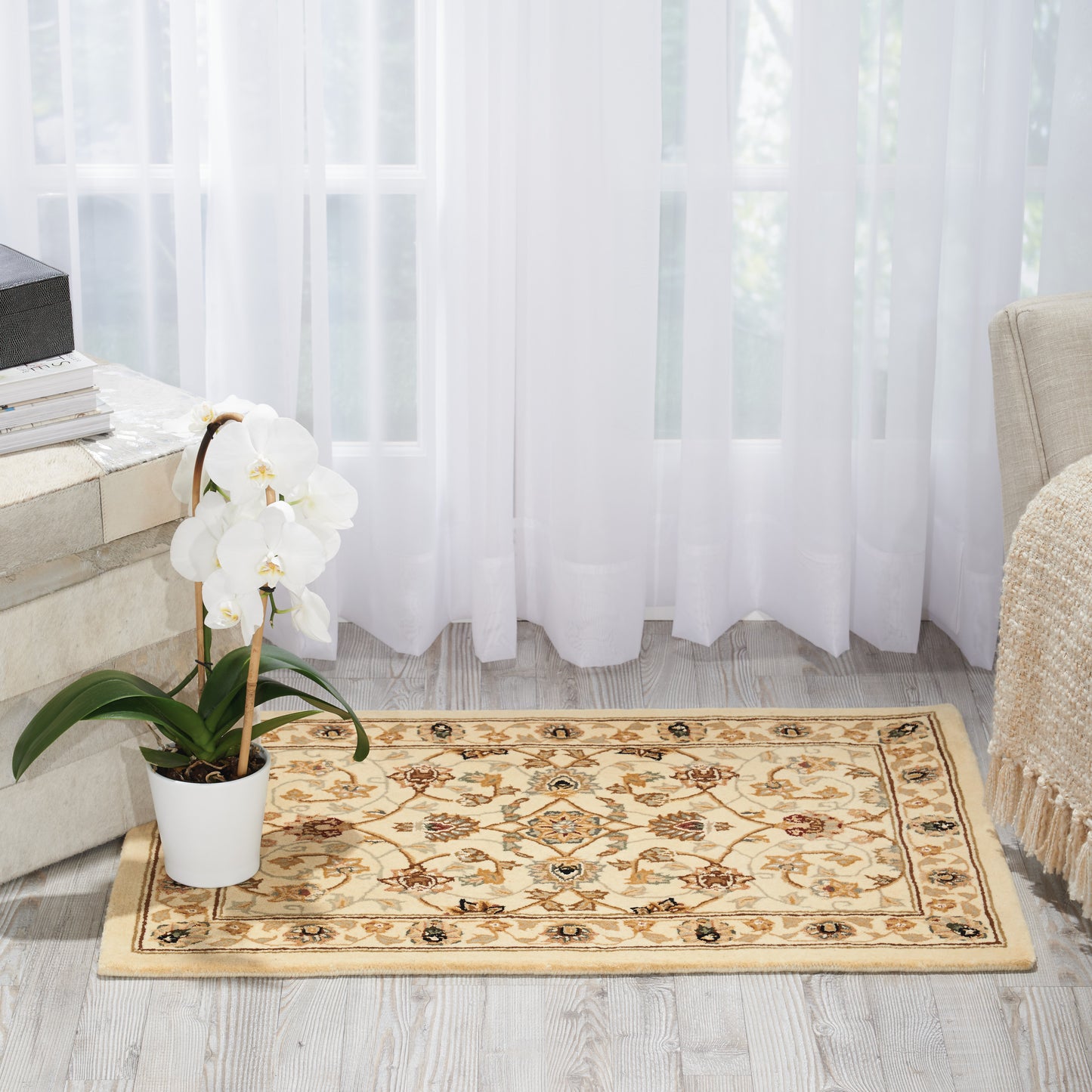 Beige oriental rug with floral medallions on a light wood floor, white orchid in a pot, books on a table, sheer white curtains.