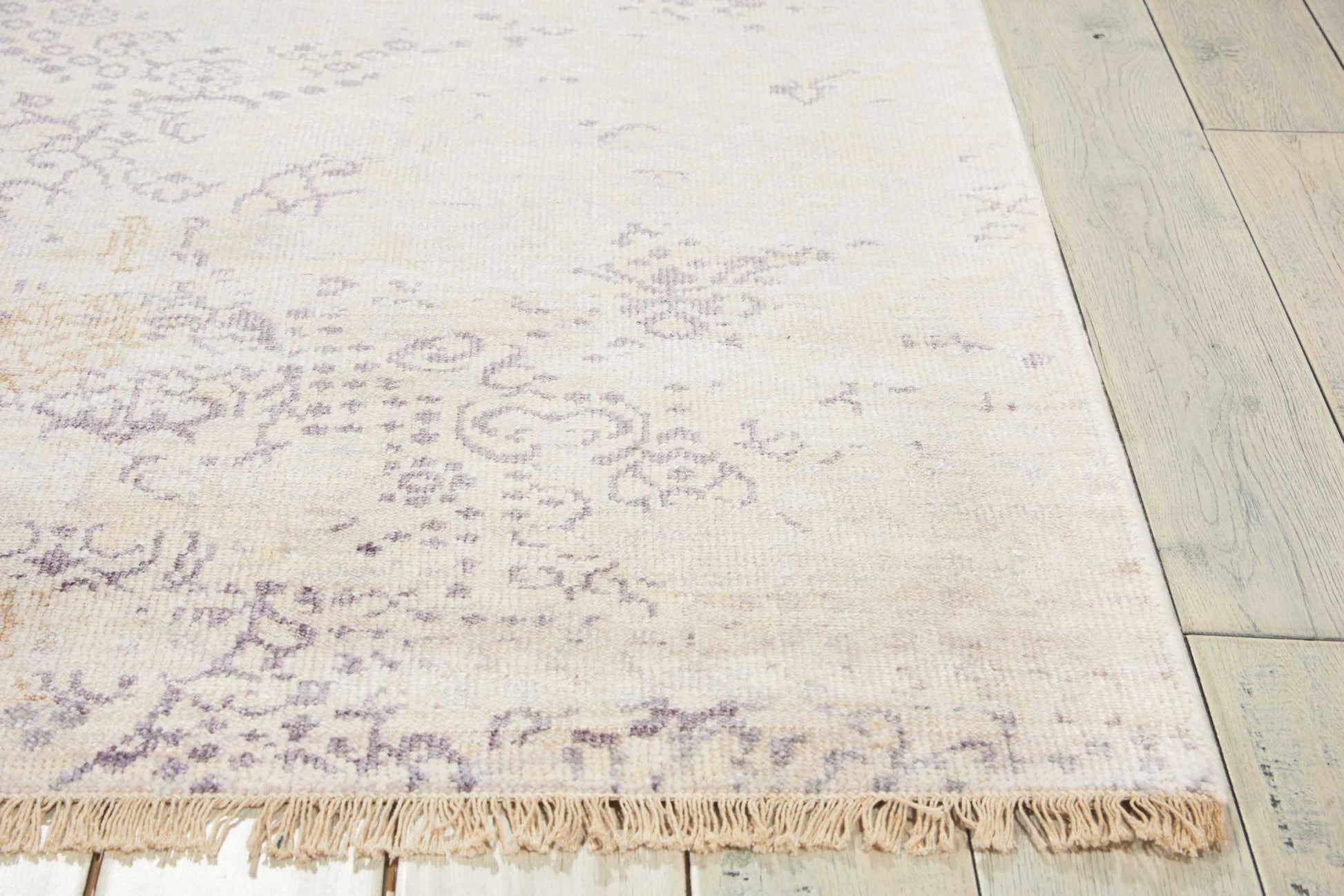 Ivory rectangular area rug with a subtle gray floral pattern and fringe tassels along the edge on a light wood floor.