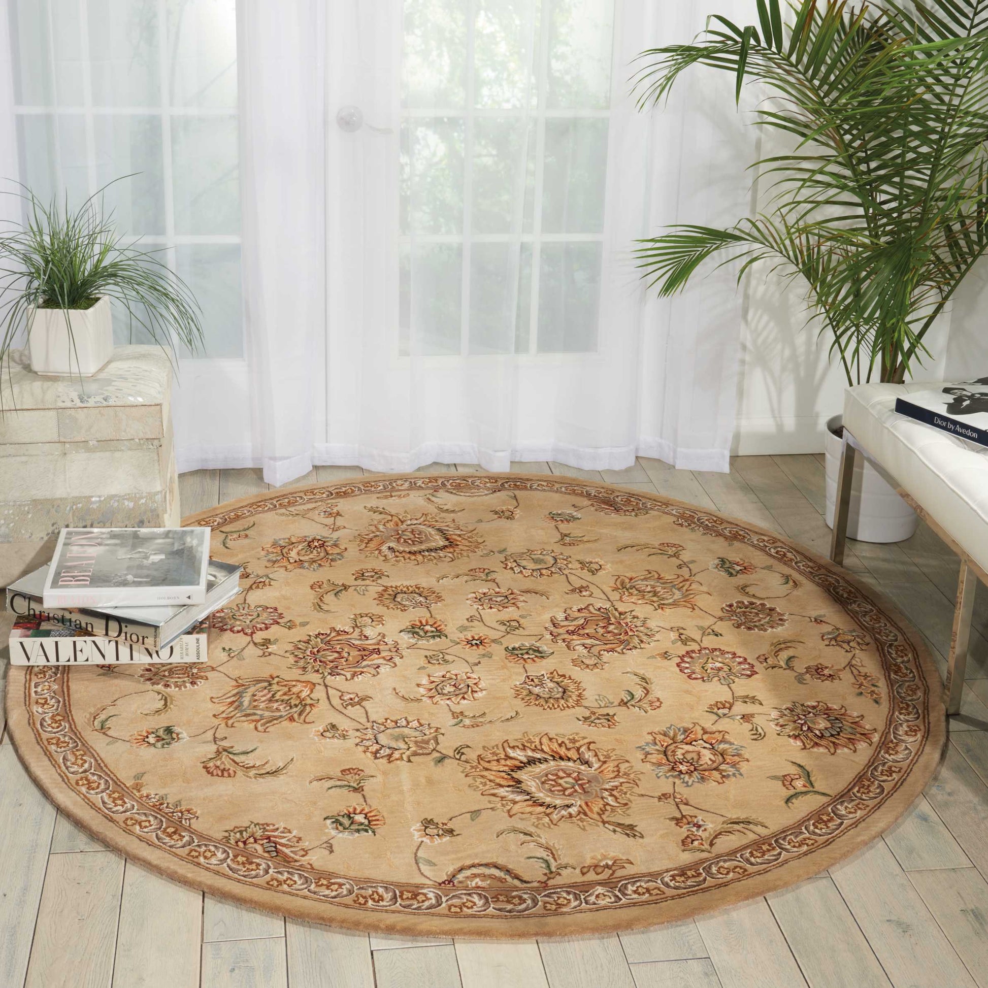 Round beige oriental rug with floral medallions and brown border on light wood floor in a bright, plant-filled living room