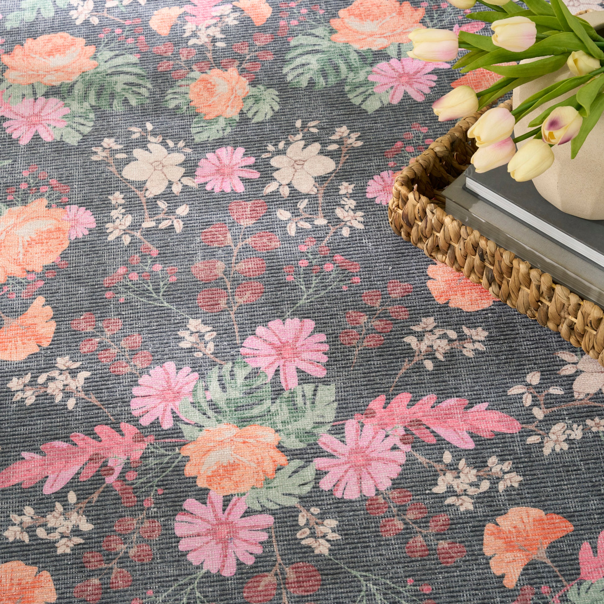 Close-up of a dark gray floral rug with pink, peach, and white flowers; woven texture, basket and tulips nearby.
