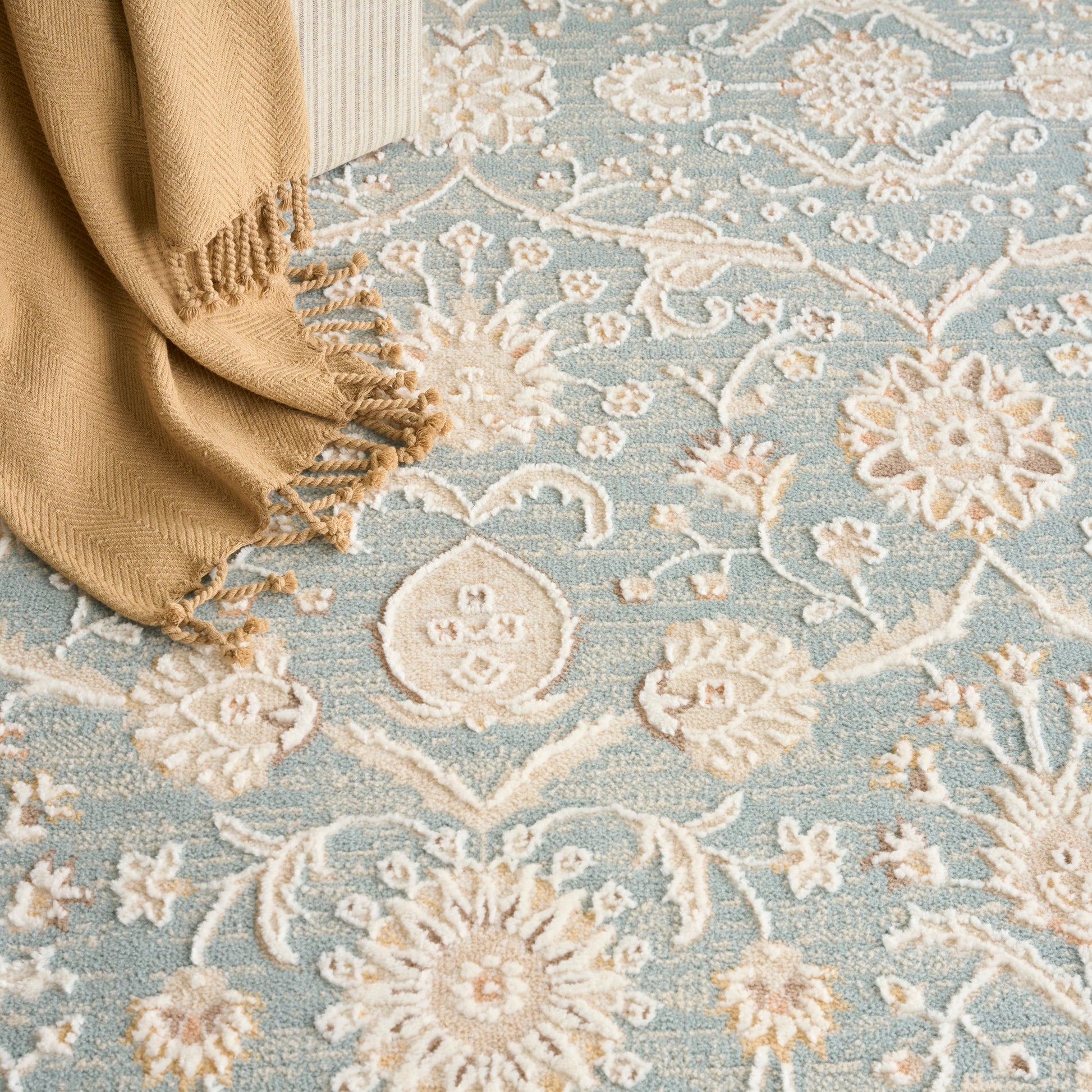 Light blue floral rug with ivory medallions and a beige fringed throw draped at the corner.