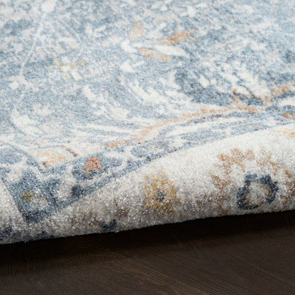 Close-up of a blue-gray textured rug with ivory and beige accents, curled edge on dark wood floor.