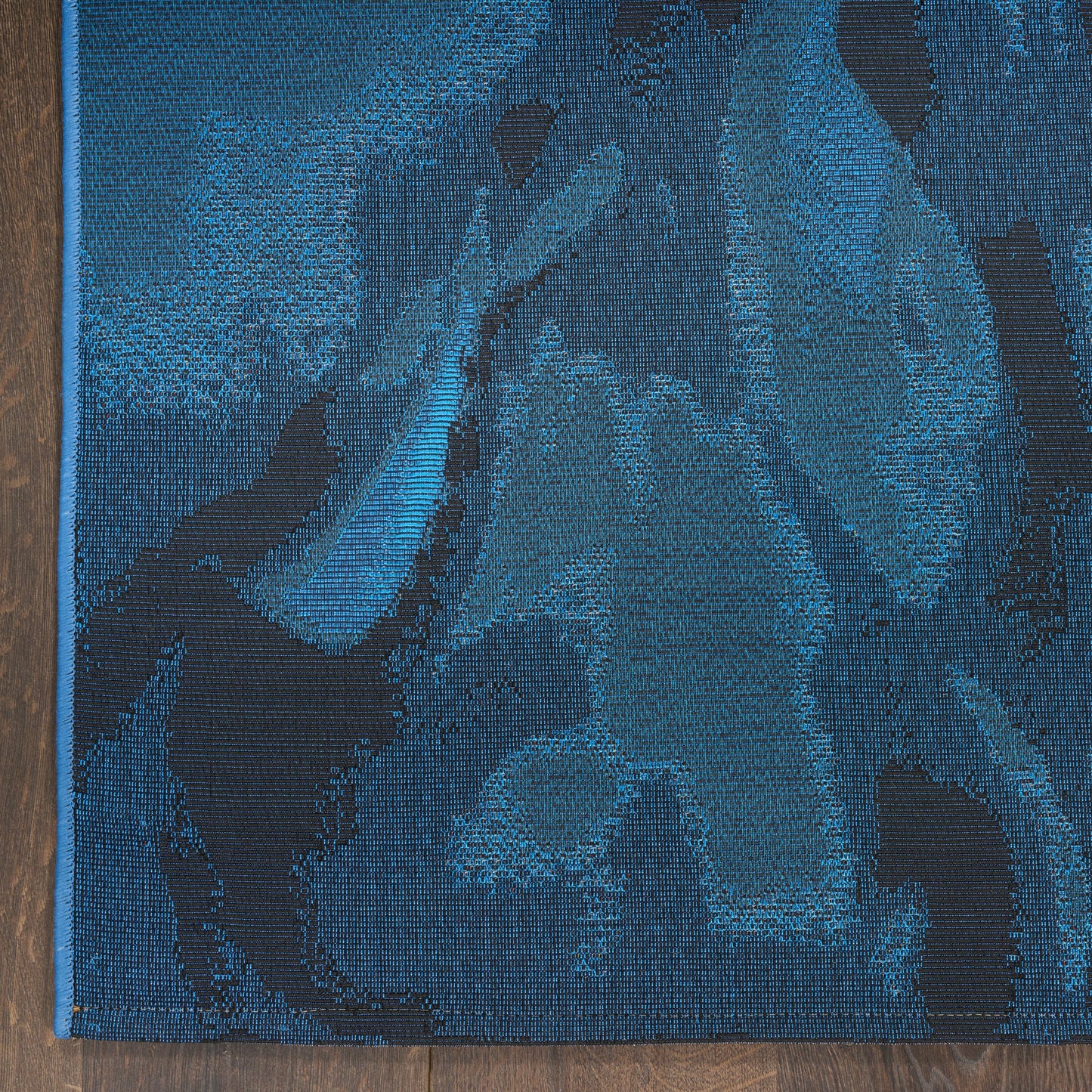 Blue woven rug with abstract floral pattern in navy and teal tones, bound edge along the left on a wooden floor.