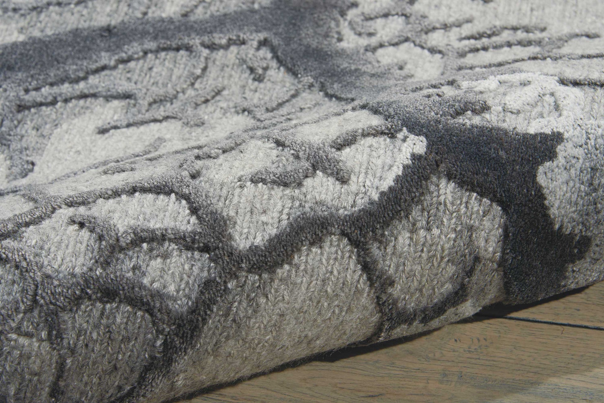 Grey and charcoal geometric patterned plush rug, rolled edge on a wooden floor.