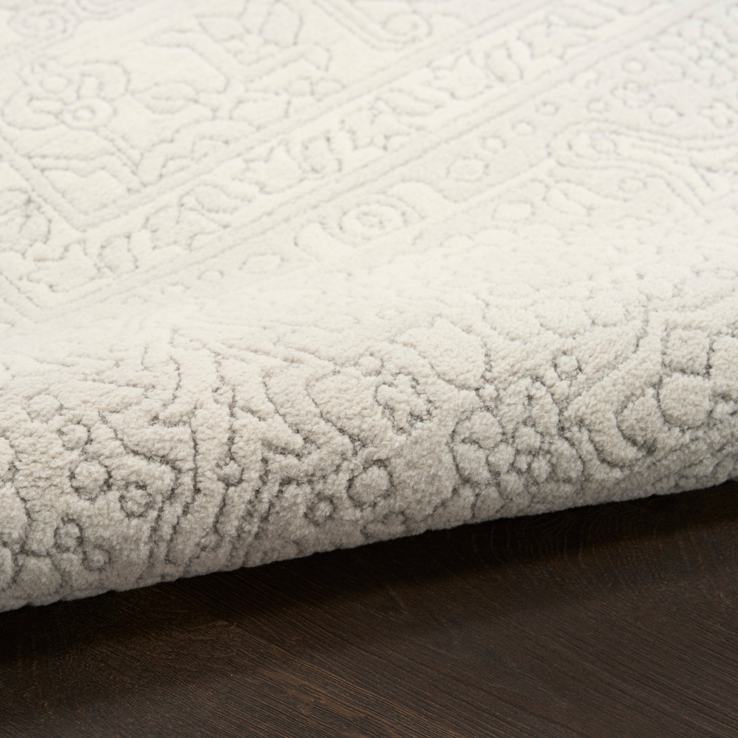 Cream textured rug with intricate embossed floral and geometric border, partially folded on dark wood floor.